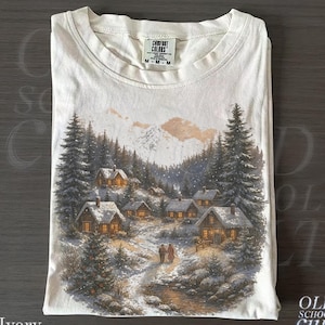 May include: Ivory-colored t-shirt featuring a winter village scene with snow-covered houses, evergreen trees, and a mountain backdrop. The shirt has a Comfort Colors tag and the text "OLD SCHOOL CULT".