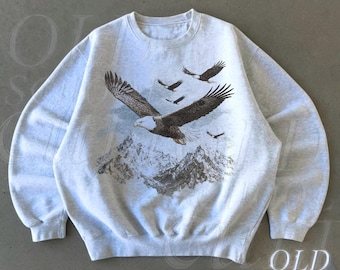 May include: Ash-colored sweatshirt featuring a detailed graphic of a bald eagle in flight above a mountain range. Several other eagles are in the sky. The text "OLD SCHOOL CULT" is printed on the lower right. The word "Ash" is printed on the lower left.