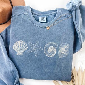Peut inclure: Sweat-shirt bleu jean à col rond et manches longues. Le devant du vêtement présente un motif blanc d'un coquillage, d'une étoile de mer et d'autres créatures marines. Le texte "Blue Jean" est en bas.