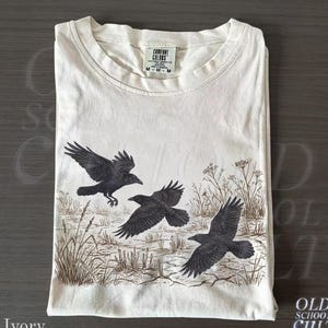 Könnte beinhalten: Elfenbeinfarbenes T-Shirt mit einer Grafik von drei schwarzen Krähen im Flug über einem Feld. Das Shirt hat einen Rundhalsausschnitt und ein Etikett mit der Aufschrift "Comfort Colors". Der Text "Old School Cult" ist sichtbar.