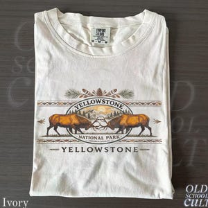 May include: Ivory-colored t-shirt featuring a Yellowstone National Park design. The graphic includes two bison, the park name, and the text "Lodging & Wildlife." The shirt is folded and has a Comfort Colors tag.