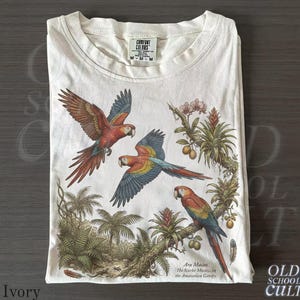 May include: Ivory t-shirt with a detailed illustration of Scarlet Macaws in a tropical setting. The design includes the text "Ara Macao, The Scarlet Macaw, in the Amazonian Canopy." The shirt is from Comfort Colors and is size M.