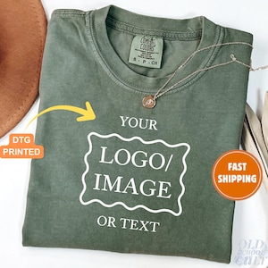 May include: An olive green t-shirt with the text "YOUR LOGO/IMAGE OR TEXT" in a white, outlined font. A gold necklace with a pendant rests on the shirt. The shirt has a Comfort Colors tag.