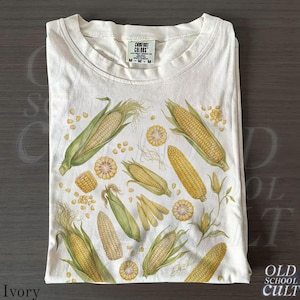 Könnte beinhalten: Elfenbeinfarbenes T-Shirt mit einer Aquarellillustration von Maiskolben, Körnern und Maisblättern. Das Shirt hat einen Rundhalsausschnitt und kurze Ärmel. Das Etikett lautet "Comfort Colors". Der Text "Old School Cult" befindet sich unten rechts.