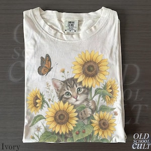 Può includere: T-shirt color avorio con grafica di un gattino tra girasoli e una farfalla. La maglietta ha girocollo e maniche corte. L'etichetta del marchio "Comfort Colors" è visibile allo scollo. La maglietta è piegata con cura.