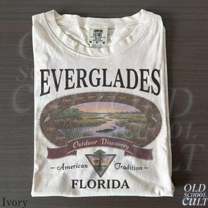 以下が含まれることがあります： 「EVERGLADES」の文字と、ワニ、鳥、夕焼けを描いた楕円形のイラストがプリントされたアイボリー色のTシャツ。「Outdoor Discovery - American Tradition - FLORIDA」と書かれたバナーが下にあります。