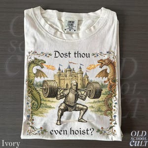 May include: Ivory t-shirt with a knight lifting weights, a castle, and dragons breathing fire. The text reads "Dost thou even hoist?" above and below the image. The shirt has a Comfort Colors tag. The shirt is folded.