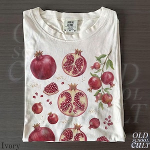 May include: Ivory-colored t-shirt featuring a watercolor design of pomegranates. The design includes whole and sliced pomegranates, seeds, and a branch with leaves. The shirt has a crew neck and short sleeves. The tag reads "COMFORT COLORS".