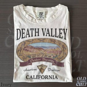 Può includere: T-shirt color avorio con grafica Death Valley. Il design include un'illustrazione ovale del paesaggio desertico, le parole "DEATH VALLEY", "Outdoor Discovery", "American Tradition" e "CALIFORNIA".