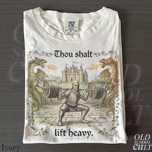 Puede incluir: Camiseta de color marfil con un diseño medieval. Muestra a un caballero levantando pesas, flanqueado por dragones, con el texto "Thou shalt lift heavy". La camiseta tiene cuello redondo y una etiqueta Comfort Colors.
