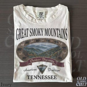 May include: Ivory-colored t-shirt featuring the text "Great Smoky Mountains" above a scenic illustration of mountains, trees, and wildlife. The shirt also includes the words "Outdoor Discovery," "American Tradition," and "Tennessee."