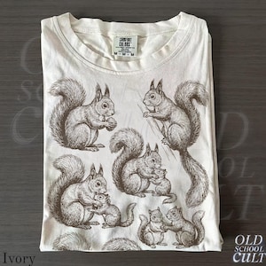 May include: Ivory-colored t-shirt featuring a detailed sepia-toned illustration of squirrels in various poses. The design includes squirrels holding nuts and interacting with each other. The shirt has a Comfort Colors tag.