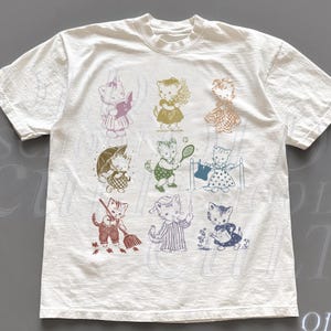 May include: White t-shirt featuring a grid of vintage-style illustrations of cats in various poses. Each cat is depicted in a different outfit and activity, with a range of colors including pink, green, and blue. The shirt has a crew neck and short sleeves.