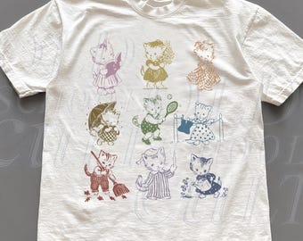 May include: White t-shirt featuring a grid of vintage-style illustrations of cats in various poses. Each cat is depicted in a different outfit and activity, with a range of colors including pink, green, and blue. The shirt has a crew neck and short sleeves.