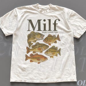 Peut inclure: T-shirt blanc avec un motif de six poissons bruns et orangés. Le mot "Milf" est imprimé au-dessus des poissons, avec "Man I Love Fish" en dessous. Le t-shirt a un col rond et des manches courtes.