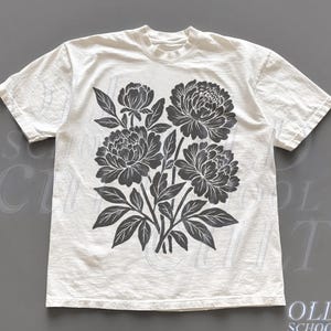 May include: Off-white t-shirt featuring a large, dark gray floral design of peonies and leaves. The short-sleeved shirt has a crew neck and a slightly textured appearance. The words "OLD SCHOOL CULT" are printed on the bottom right corner.