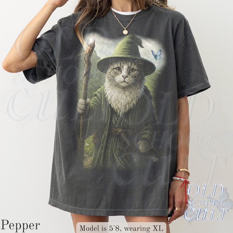 May include: Dark gray t-shirt featuring a whimsical illustration of a cat dressed as a wizard, holding a staff. The cat wears a green hat and robe, with a blue butterfly in the background. The text "OLD SCHOOL CULT" is visible on the shirt.
