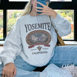 May include: A light gray sweatshirt with the word "YOSEMITE" in dark brown lettering. Below the word is an oval graphic with a mountain scene and the words "Outdoor Discovery". The sweatshirt also has the words "American Tradition CALIFORNIA".