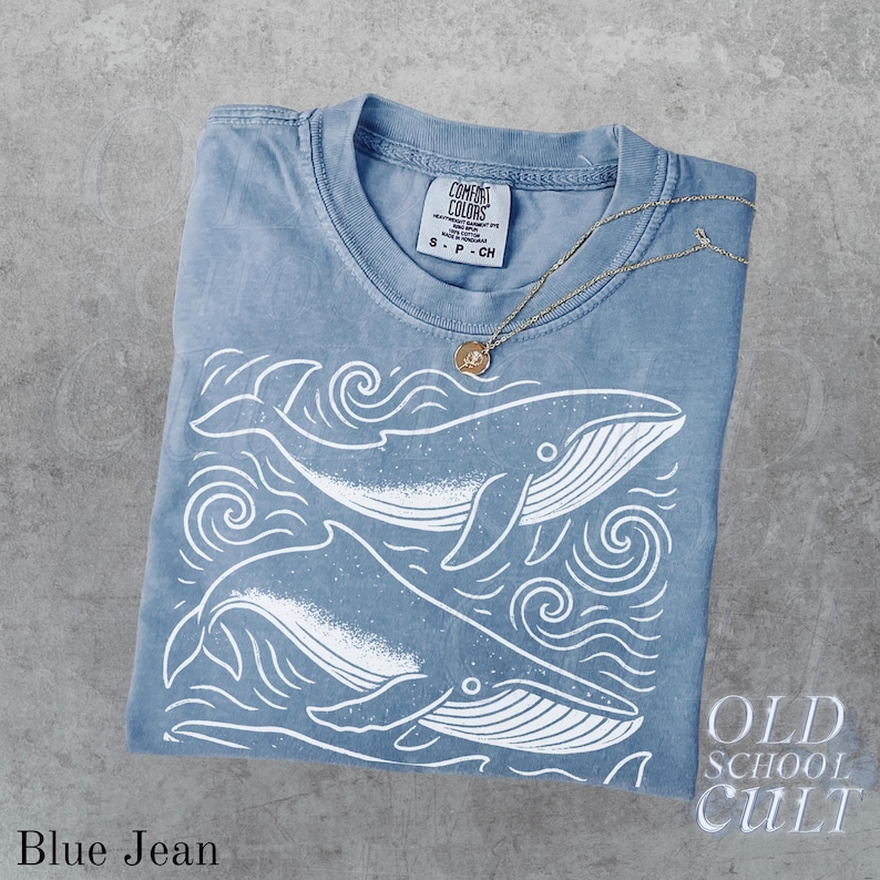 May include: A blue jean Comfort Colors t-shirt with a white whale design and a gold necklace. The shirt features two whales with wave details. The tag reads "Comfort Colors" and "S-P-CH". The words "OLD SCHOOL CULT" are visible.