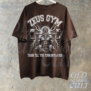 May include: Brown t-shirt with a white graphic of Zeus lifting weights. The graphic includes the text "Zeus Gym" and "Train till you turn into a god".