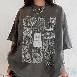 May include: Dark grey t-shirt featuring a grid of white cat illustrations. The cats are depicted in various poses, including sitting, lying down, and curled up. The shirt has a vintage, washed-out look. The text "OLD SCHOOL CULT" is visible on the bottom right.