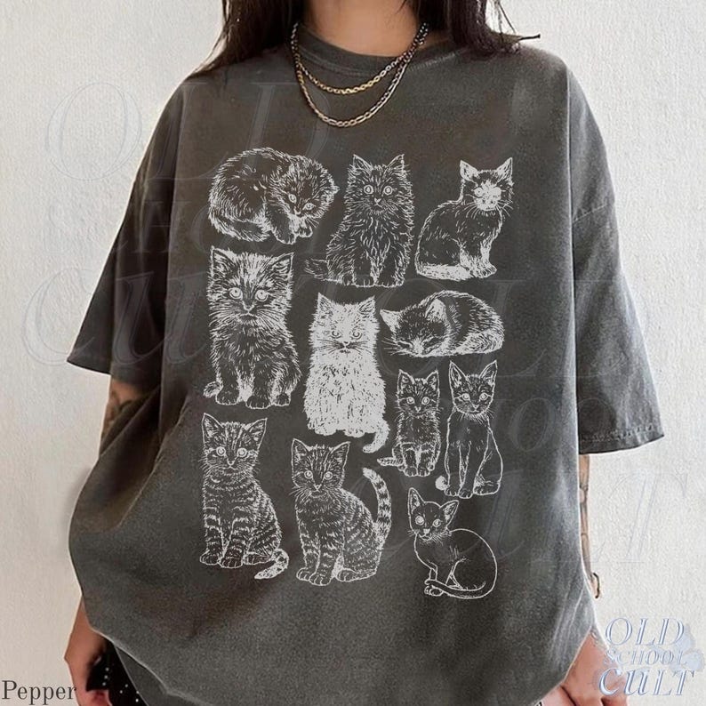 May include: Dark grey t-shirt featuring a grid of white cat illustrations. The cats are depicted in various poses, including sitting, lying down, and curled up. The shirt has a vintage, washed-out look. The text "OLD SCHOOL CULT" is visible on the bottom right.