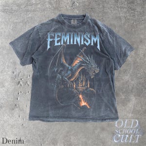 May include: A denim-colored t-shirt featuring the word "FEMINISM" in a stylized font above a detailed illustration of a dragon perched in front of a castle. The dragon is blue and black, with orange flames below the castle.