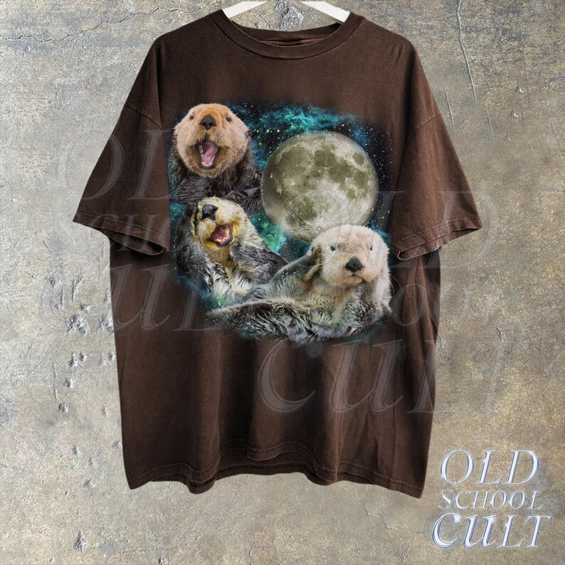 May include: A brown t-shirt with a graphic print of three sea otters swimming in a galaxy with a full moon. The otters are brown and white with black eyes and noses.
