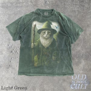 May include: Light green t-shirt featuring a whimsical illustration of a cat dressed as a wizard, complete with a hat and staff. The cat is depicted with a long white beard and is set against a soft, cloudy background. The shirt also has the text "OLD SCHOOL CULT".