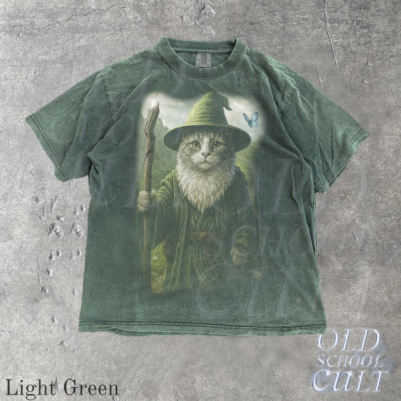 May include: Light green t-shirt featuring a whimsical illustration of a cat dressed as a wizard, complete with a hat and staff. The cat is depicted with a long white beard and is set against a soft, cloudy background. The shirt also has the text "OLD SCHOOL CULT".