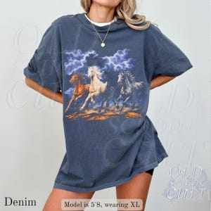 May include: Denim blue t-shirt featuring a graphic of three running horses under a stormy sky. The horses are in shades of brown, white, and gray. The shirt has short sleeves and a relaxed fit. The text "OLD SCHOOL CULT" is visible in the background.