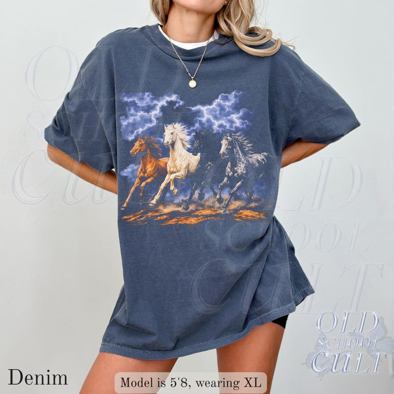 May include: Denim blue t-shirt featuring a graphic of three running horses under a stormy sky. The horses are in shades of brown, white, and gray. The shirt has short sleeves and a relaxed fit. The text "OLD SCHOOL CULT" is visible in the background.