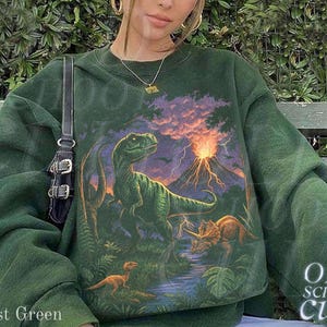 Op de afbeelding: Bostgroen sweatshirt met een dinosaurusafbeelding, een vulkaan en andere dinosaurussen. Het sweatshirt heeft de tekst "OLD SCHOOL CULT" rechtsonder. De woorden "Forest Green" staan linksonder.