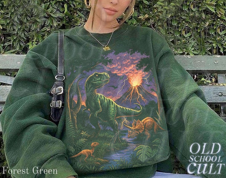 Op de afbeelding: Bostgroen sweatshirt met een dinosaurusafbeelding, een vulkaan en andere dinosaurussen. Het sweatshirt heeft de tekst "OLD SCHOOL CULT" rechtsonder. De woorden "Forest Green" staan linksonder.