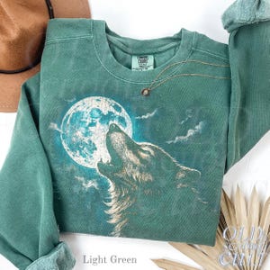 May include: Light green sweatshirt featuring a howling wolf graphic against a full moon. The sweatshirt has a crew neck and long sleeves. A gold necklace with a small pendant is draped on the shirt. The text "OLD SCHOOL CHILL" is visible.