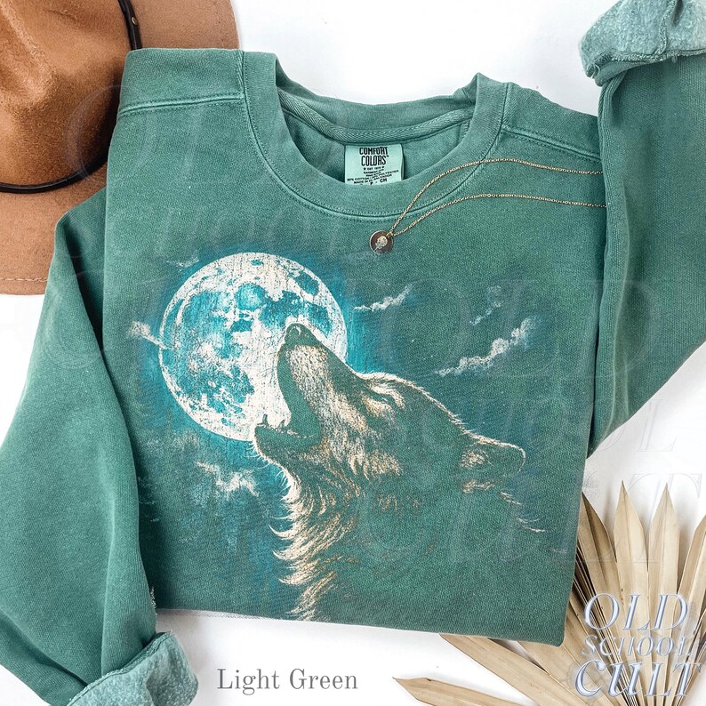May include: Light green sweatshirt featuring a howling wolf graphic against a full moon. The sweatshirt has a crew neck and long sleeves. A gold necklace with a small pendant is draped on the shirt. The text "OLD SCHOOL CHILL" is visible.