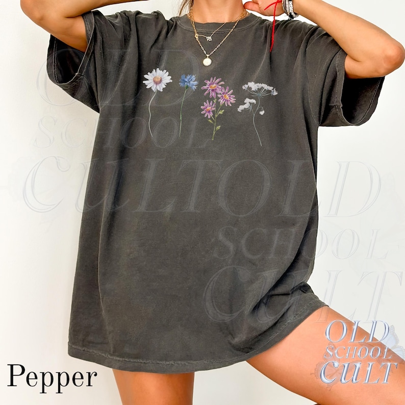 Pu&ograve; includere: Una maglietta grigio scuro con un design floreale con fiori bianchi, blu e viola. La maglietta ha un aspetto vintage e sbiadito e la parola "Pepper" &egrave; stampata in basso a sinistra. Parte della collezione "Old School Cult".