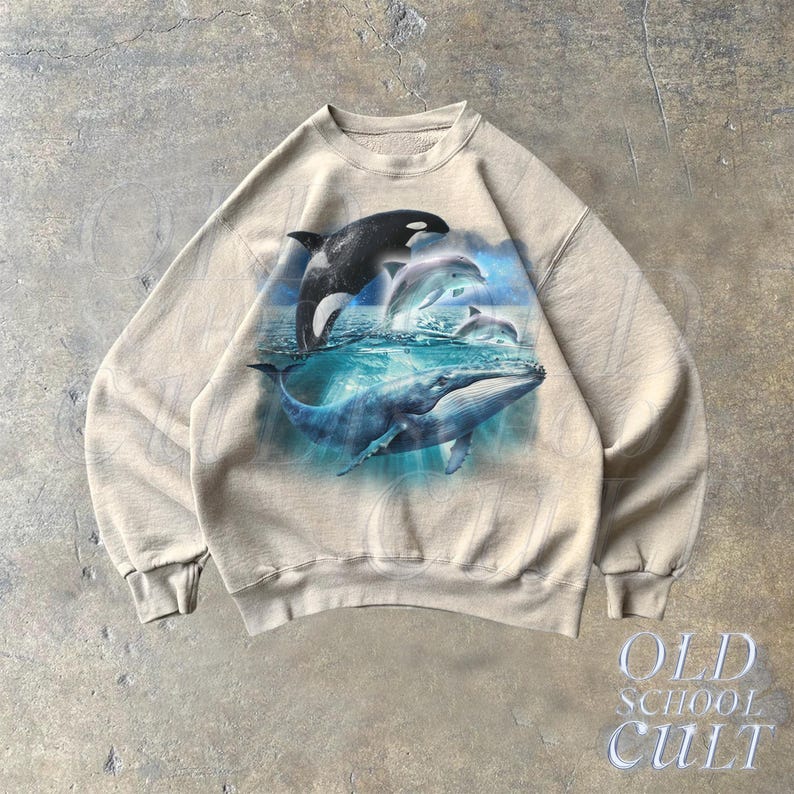 May include: A beige sweatshirt with a graphic print of a whale and dolphins swimming in the ocean. The graphic is in shades of blue, black, and white.