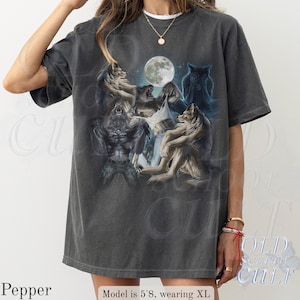 May include: Dark gray t-shirt featuring a graphic of wolves and a werewolf under a full moon. The design includes various wolf poses and a detailed illustration style. The shirt has a vintage, worn look.