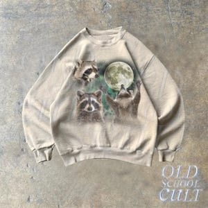 Op de afbeelding: Een beige sweatshirt met een grafische print van drie wasberen en een volle maan. De wasberen kijken naar de maan.