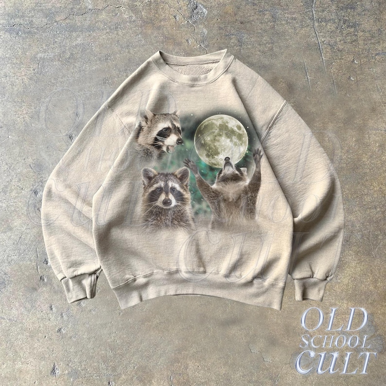 Op de afbeelding: Een beige sweatshirt met een grafische print van drie wasberen en een volle maan. De wasberen kijken naar de maan.