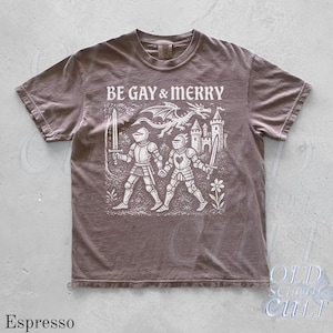 Puede incluir: Camiseta color espresso con el texto "BE GAY & MERRY" sobre un gr&aacute;fico de dos caballeros, un drag&oacute;n y un castillo. La camiseta tiene cuello redondo y mangas cortas. La marca "OLD SCHOOL CULT" est&aacute; impresa en la parte inferior derecha.