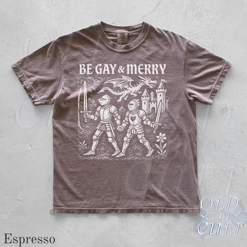 Puede incluir: Camiseta color espresso con el texto "BE GAY & MERRY" sobre un gr&aacute;fico de dos caballeros, un drag&oacute;n y un castillo. La camiseta tiene cuello redondo y mangas cortas. La marca "OLD SCHOOL CULT" est&aacute; impresa en la parte inferior derecha.