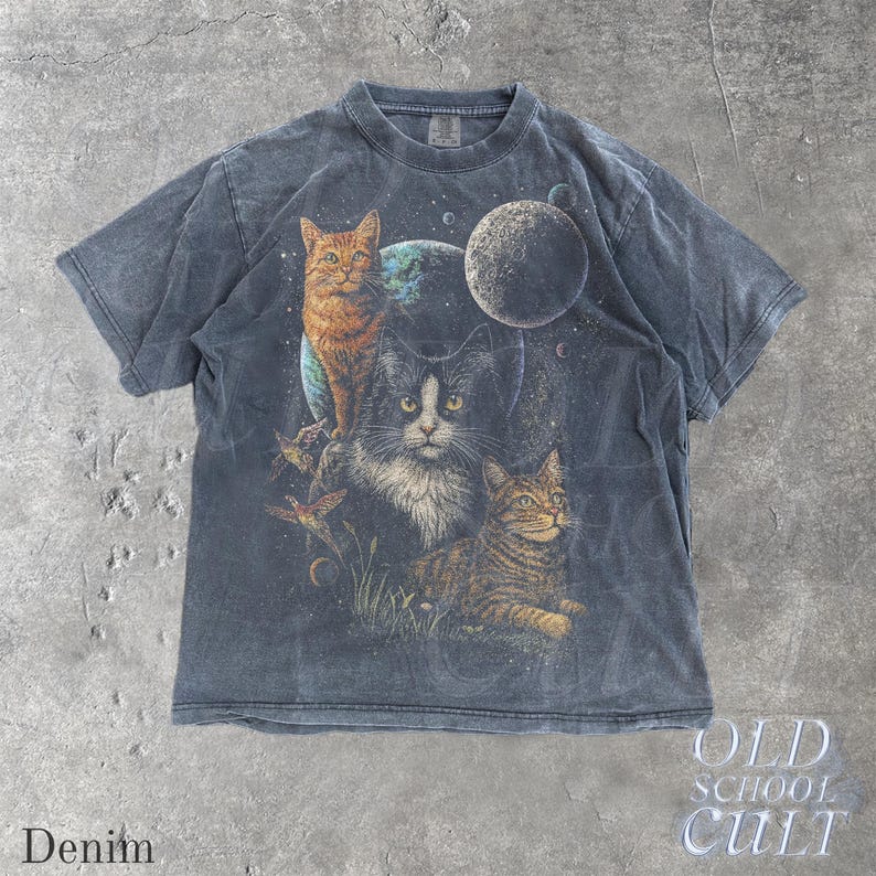 以下が含まれることがあります： デニムカラーのTシャツ。3匹の猫と惑星を背景にしたグラフィック。猫はオレンジ、白黒、茶色のトラ柄。シャツには「OLD SCHOOL CULT」と「Denim」の文字が入っています。