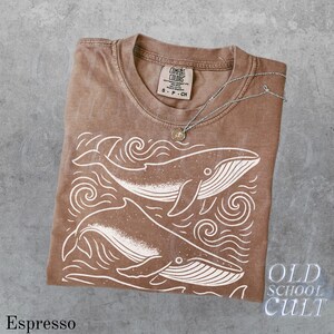 May include: Espresso-colored t-shirt with a white whale graphic and the text "OLD SCHOOL CULT". The shirt is folded and has a Comfort Colors tag. A silver necklace with a small pendant rests on the shirt.