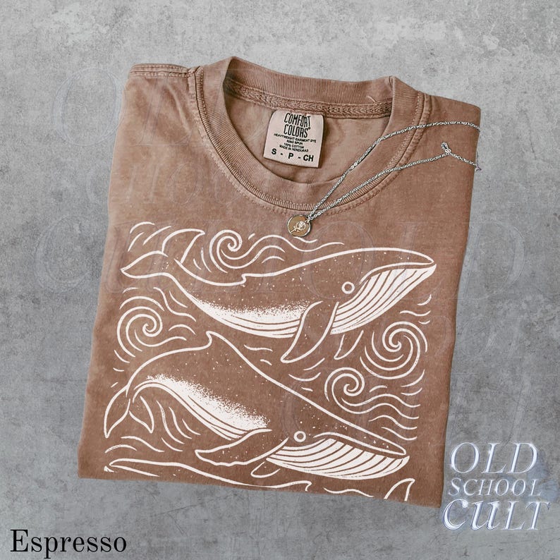 May include: Espresso-colored t-shirt with a white whale graphic and the text "OLD SCHOOL CULT". The shirt is folded and has a Comfort Colors tag. A silver necklace with a small pendant rests on the shirt.
