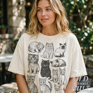 May include: Cream-colored t-shirt featuring a collection of black and white cat illustrations. The cats are depicted in various poses, including sitting, sleeping, and standing. The text "OLD SCHOOL CULT" is printed in a stylized font on the lower right.