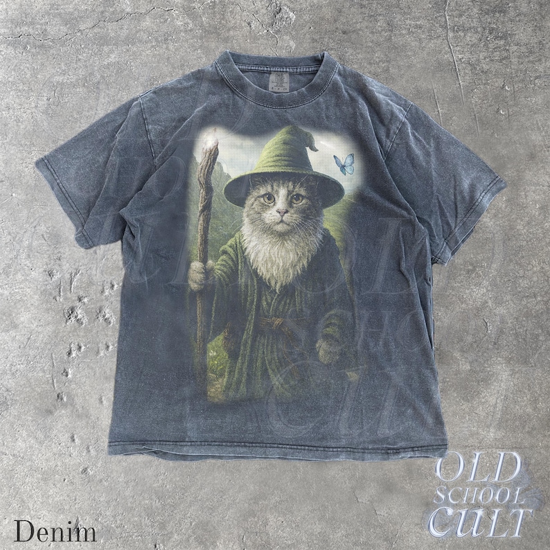 May include: Denim-colored t-shirt featuring a whimsical illustration of a cat dressed as a wizard, complete with a hat, staff, and flowing robes. A blue butterfly is also present in the design. The words "Old School Cult" and "Denim" are printed on the shirt.