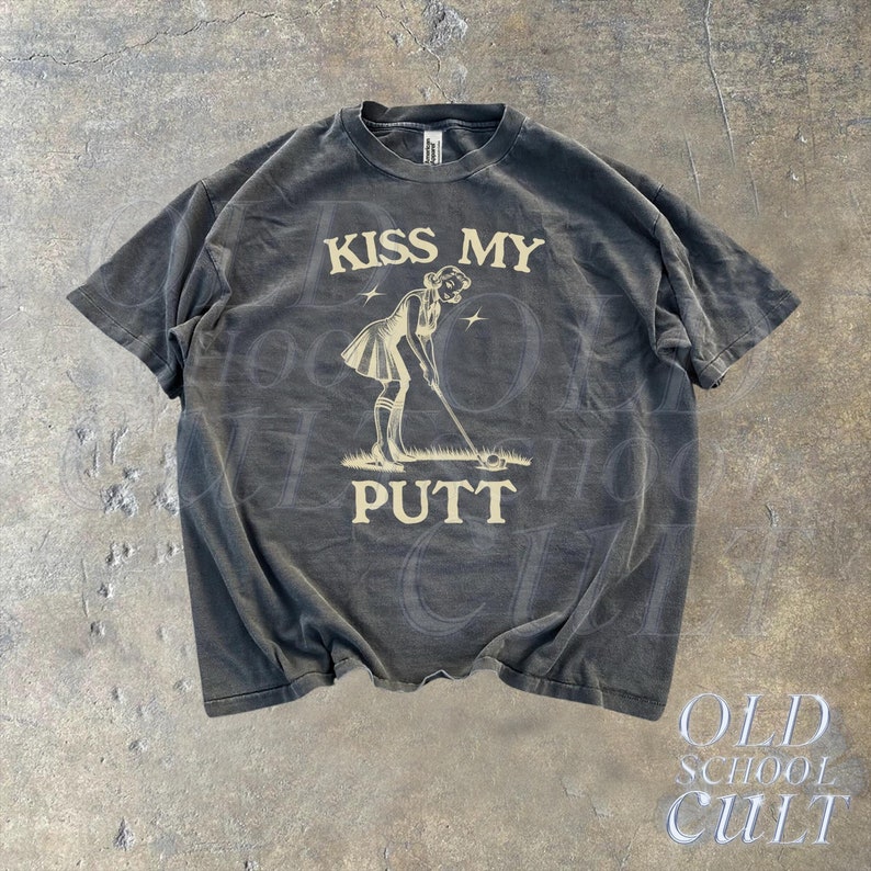 May include: A black t-shirt with a vintage-style graphic of a woman putting a golf ball. The graphic is in a cream color and says "Kiss My Putt".