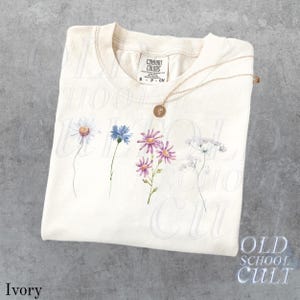 Pu&ograve; includere: T-shirt color avorio con un design floreale ad acquerello con margherite, fiordalisi e altri fiori di campo. Una collana d'oro con un ciondolo &egrave; appoggiata sulla maglietta. La maglietta ha un'etichetta Comfort Colors.